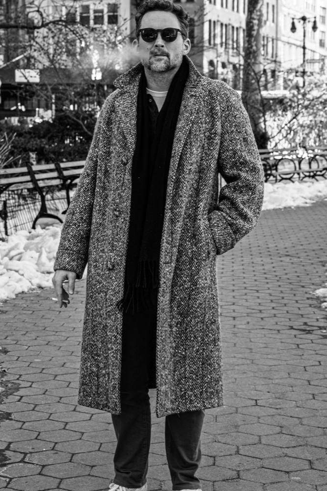 Jesse Sherwood standing on a snowy park path in the city, wearing sunglasses, a tweed coat, and a scarf.