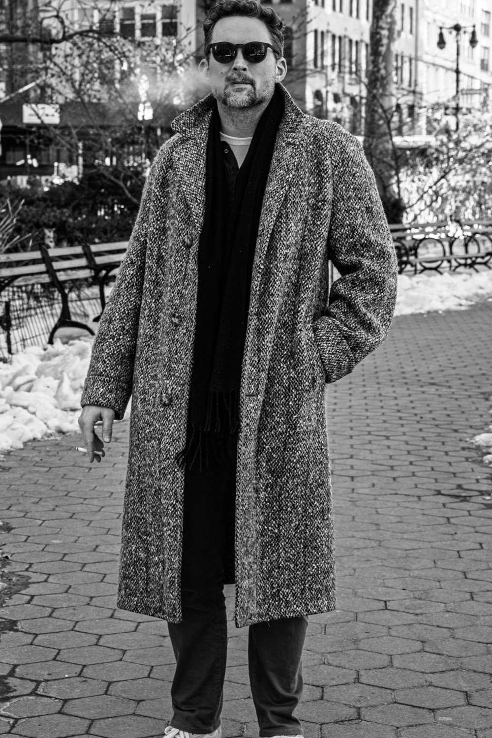 Jesse Sherwood standing on a snowy park path in the city, wearing sunglasses, a tweed coat, and a scarf.