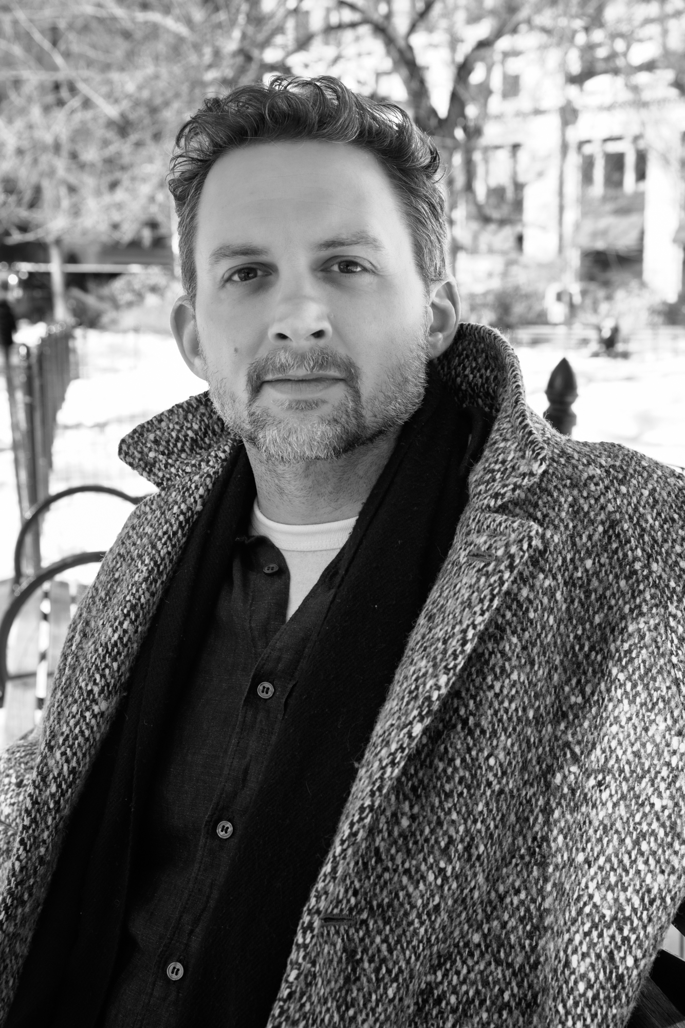 Jesse Sherwood on a bench in a park in the city wearing a large tweed coat.